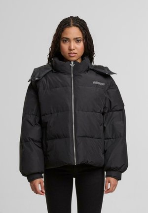 Ung kvinna med flätat hår bär en svart pufferjacka med hög krage och dragkedja framtill, stående mot en enfärgad ljusgrå bakgrund.