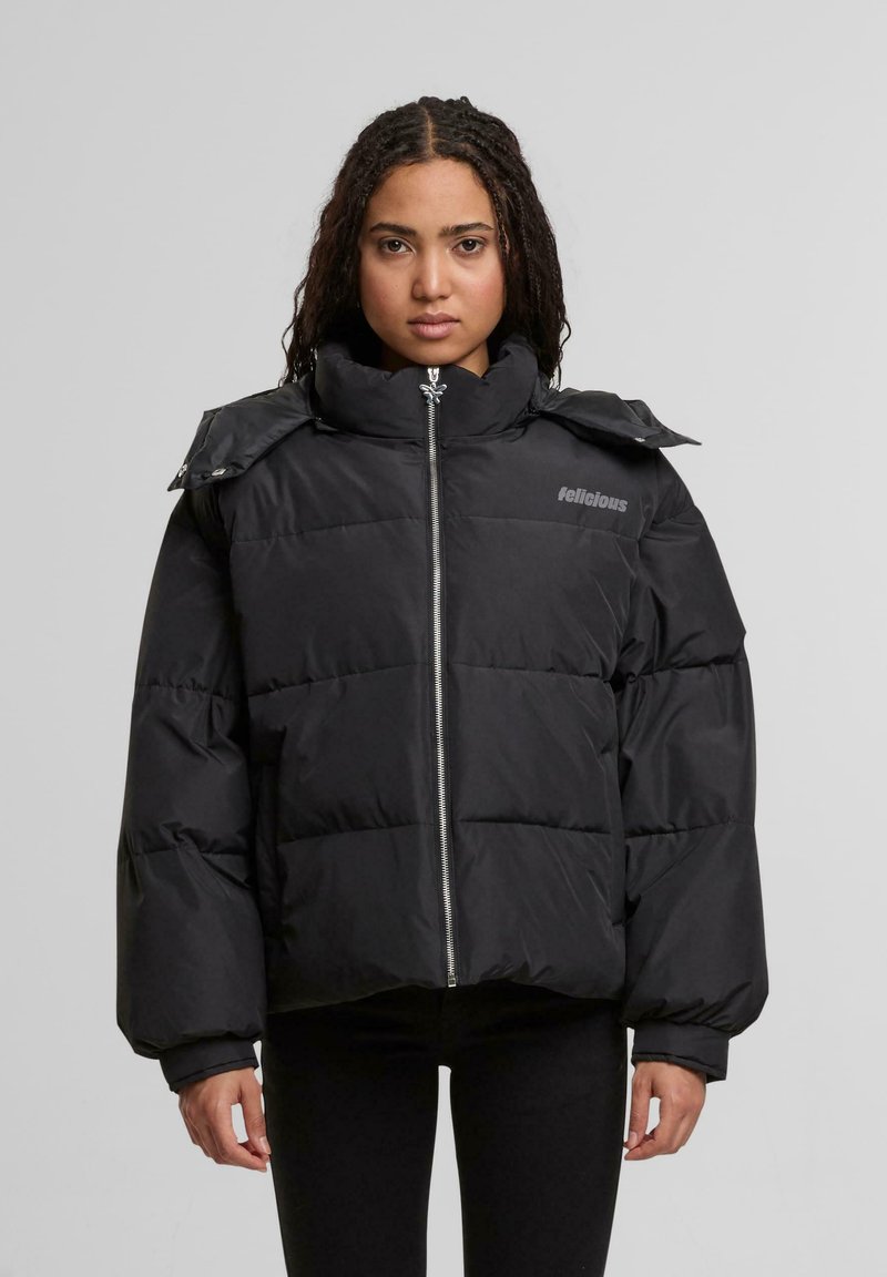 Giovane donna con capelli intrecciati che indossa un giubbotto trapuntato nero con colletto alto e zip frontale, in piedi di fronte a uno sfondo grigio chiaro uniformemente.