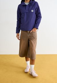 Persona vestida con una chaqueta con capucha morada, pantalones cortos marrones hasta la rodilla, calcetines blancos y zapatillas rosa claro sobre un suelo amarillo.