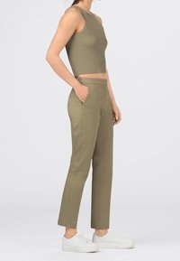 Žena v olivově zeleném bezrukávovém ribbed crop topu, s odpovídajícími tailored kalhotami a bílými teniskami, stojící s jednou rukou v kapse.