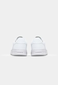 White leather sneakers with a smooth finish, featuring a low-top design, rounded edges, and a textured sole. "Lacoste" branding on the heel.
