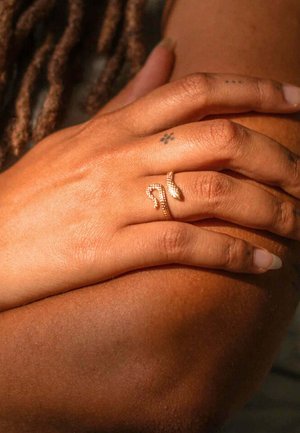 Anello a forma di serpente d'oro su un dito, con una superficie metallica liscia. La mano si appoggia su una pelle di tono più scuro, adornata da vari piccoli tatuaggi.