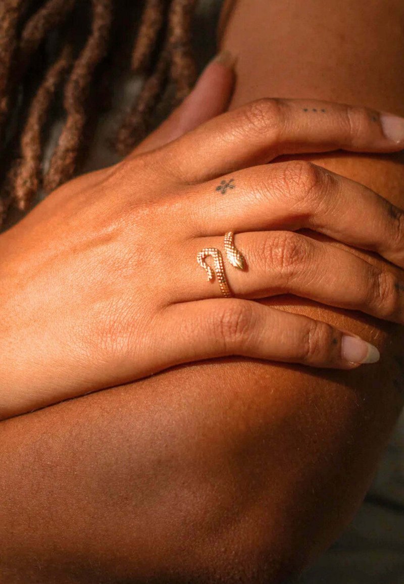Anello a forma di serpente d'oro su un dito, con una superficie metallica liscia. La mano si appoggia su una pelle di tono più scuro, adornata da vari piccoli tatuaggi.