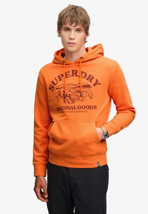 Jeune homme portant un sweat à capuche orange avec le texte et le graphisme "Superdry Original Goods", pantalon noir et montre-bracelet, debout devant un fond blanc.