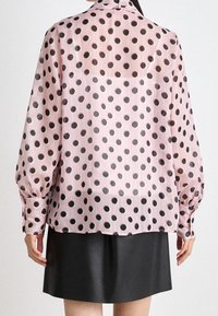 Blouse rose translucide avec de grands pois noirs, manches longues et bouffantes, et un col rond, associée à une jupe noire. Tissu texturé visible.