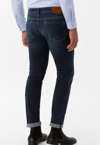 Tmavě modré, přiléhavé džíny s lesklým povrchem a koženou nášivkou na zadním pase. Srolované manžety nad elegantními černými kotníkovými botami.