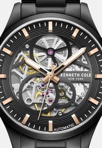 Orologio automatico nero con quadrante scheletrato trasparente. Presenta accenti in oro rosa, indicatori e un intricato movimento meccanico in giallo e argento.