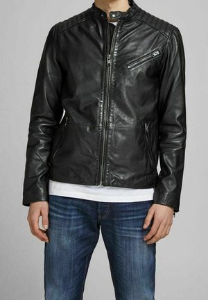 Mężczyzna w czarnej skórzanej kurtce motocyklowej z pikowanymi ramionami, przednim zamkiem błyskawicznym, zapinaną na zamek kieszenią na piersi oraz w niebieskich dżinsach.