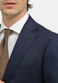 Marinblå kavaj med strukturerade axlar, slagkragar och smal passform. Matchas med en vit skjorta och ett brunt prickigt slips.