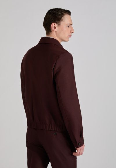 Homme portant une veste de costume bordeaux foncé et un pantalon assorti, debout de profil contre un fond gris clair uni.
