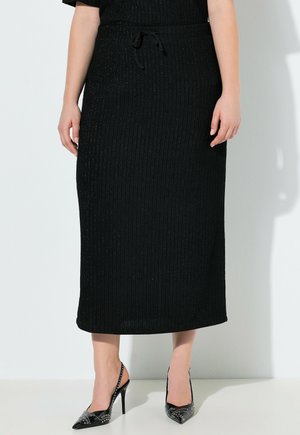 Frau trägt einen gerippten schwarzen Midirock mit Tunnelzugbund und schwarze spitze High Heels mit Nieten, steht vor einer weißen Wand.