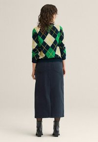 Kiharatukkainen henkilö, joka pukeutunut vihreään, tummansiniseen ja kermaiseen ruutuneuleeseen, tummaan midihameeseen, mustiin sukkiin ja mustiin korollisiin nilkkureihin, seisomassa.