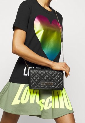 Bolso de cuero negro acolchado con una correa de cadena, que presenta una placa con el logo. El modelo lleva una camiseta negra con un gráfico de un corazón colorido y una falda.