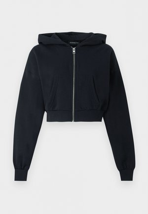 Sort cropped hættetrøje med lynlås foran, kengurulomme, lange ærmer og ribkant ved ærmeenderne på ensfarvet baggrund.