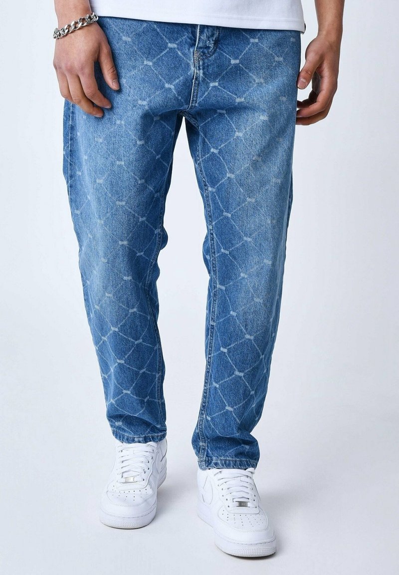 Personne portant un jean bleu avec un motif losange délavé et des baskets blanches, debout devant un fond clair uni.
