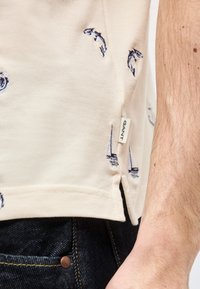 Gros plan sur l'ourlet d'une chemise beige avec des motifs brodés de poissons bleus et de voilier, une étiquette GANT, un jean en denim foncé, et l'avant-bras d'un homme.