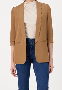 Femme portant un blazer camel aux revers festonnés, un haut beige, et un jean bleu taille haute avec une fermeture à double bouton.