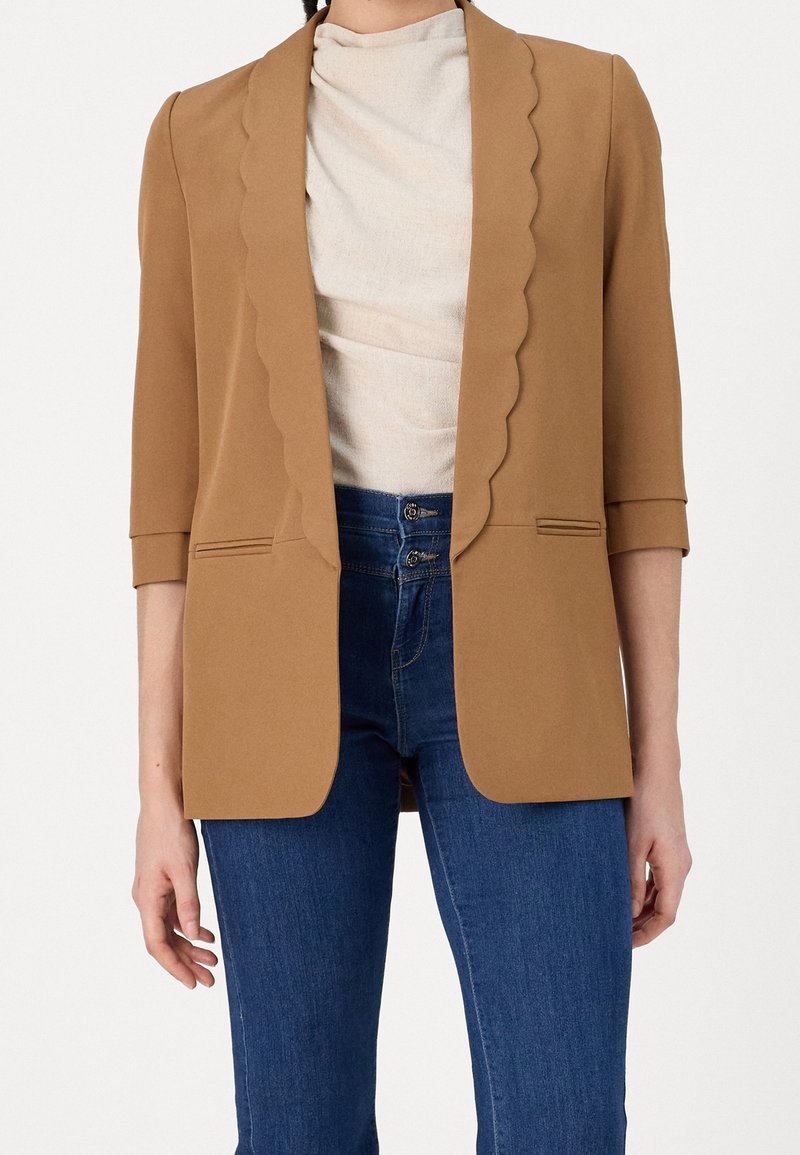 Femme portant un blazer camel aux revers festonnés, un haut beige, et un jean bleu taille haute avec une fermeture à double bouton.