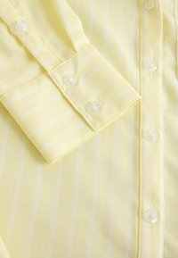 Dzeltena svītraina krekls ar gludu faktūru, ar pogas aizdares apkakli un baltām pogām priekšpusē un aprocē.