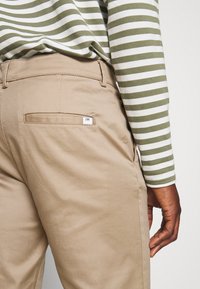 Högra sidan av en person som bär beige byxor och en långärmad skjorta med gröna och vita ränder, hand avslappnat vid sidan.