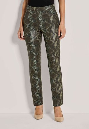 Vrouw in strakke, enkel-lange slangenprint broek en beige hoge hakken, staande tegen een effen lichte achtergrond.