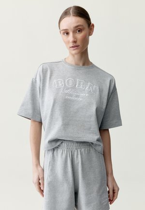 Žena nosiace sivé oversized tričko s textom „BORN Athleisure culture“ a zodpovedajúce elastické kraťasy na jednoliatom pozadí.