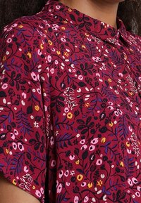 Rød blomsterbluse med krave, korte ærmer og et mønster af pink, gule og sorte blomster med grønne blade på et tekstureret stof.