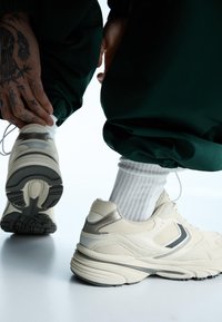 Beige Sportschuhe mit grauen Akzenten, strukturiertem Mesh und einer dicken Sohle. Weiße Sportsocken und grüne Jogginghosen sind teilweise sichtbar.