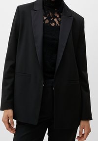 Blazer nero su misura con revers in satin, due tasche anteriori e pantaloni abbinati. Top in pizzo nero testurizzato visibile.