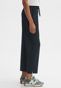 Pantalon noir à taille élastique avec fentes latérales ; tissu léger ; associé à des baskets vertes et blanches avec une semelle texturée et des lacets.
