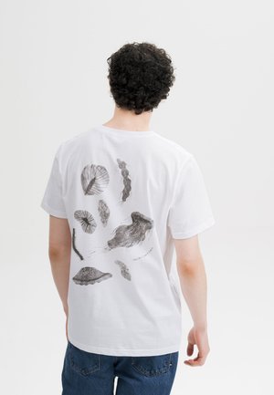 Persoon met krullend donker haar die een wit T-shirt draagt met zwarte abstracte illustraties van zeeleven op de rug, gecombineerd met een blauwe spijkerbroek.