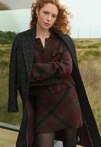 Schwarz gefleckter Mantel über einem grün-burgunder-karierten Strickoutfit, mit Knöpfen und einer Tasche. Glatte Textur, geschichteter Look.
