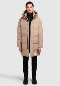 Abrigo tipo puffer beige con capucha, con un patrón acolchado, botones a presión en la parte delantera y una etiqueta de marca visible en el dobladillo. Lleva puesto un suéter de cuello alto negro.