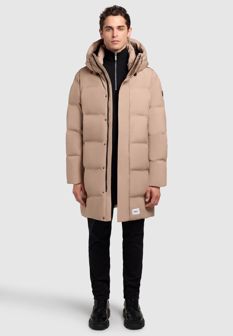 Abrigo tipo puffer beige con capucha, con un patrón acolchado, botones a presión en la parte delantera y una etiqueta de marca visible en el dobladillo. Lleva puesto un suéter de cuello alto negro.