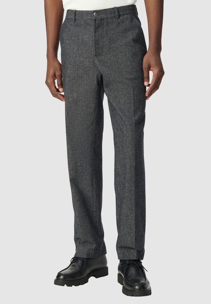 Pantaloni in misto lana grigio scuro con taglio dritto, passanti per la cintura, tasche anteriori e chiusura a bottone. Indossati con scarpe nere stringate.