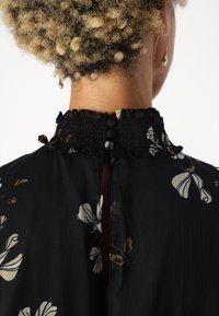 Kiharinen henkilö wear musta kukallinen blusa, jossa on rypytetty korkea kaulus, kiinnitettynä kolmella kankaasta päällystetyllä nappilla takana.