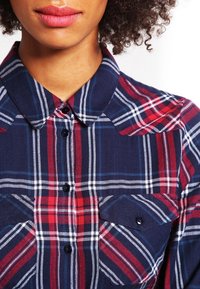 Camicia con bottoni, caratterizzata da un motivo a quadri blu navy, rosso e bianco. Presenta un colletto appuntito e due tasche sul petto con bottoni neri.