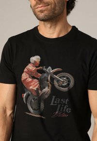 Man in een zwart T-shirt met de afbeelding van een oudere vrouw in een roze outfit die een motorfiets wheelie maakt, met de tekst "Last Life Deluxe."