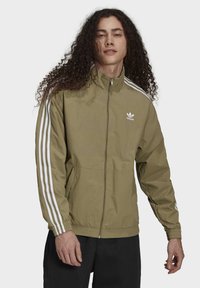 Olivgrön zip-up jacka med hög krage, med vita tre-stripes accenter längs ärmarna och en liten vit logotyp på bröstet.