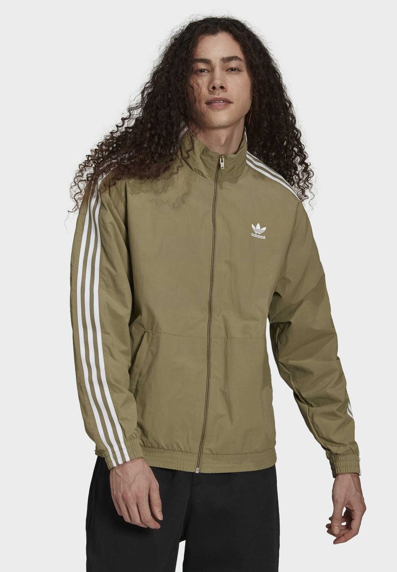 Olivgrön zip-up jacka med hög krage, med vita tre-stripes accenter längs ärmarna och en liten vit logotyp på bröstet.