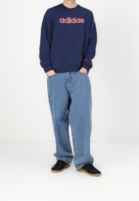 Person som bär en marinblå Adidas-hoodie med röd logotyp, lösa ljusblå jeans och svarta sneakers som står framför en enkel vit bakgrund.