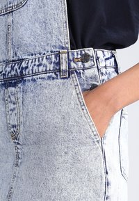 Person som bär acid-wash denimoverall med hand i framfickan, i kombination med en svart skjorta.