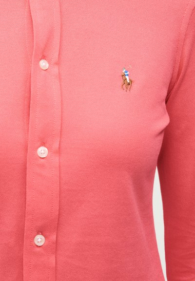 Korallszínű, texturált, gombos ing, amelyen a mellrészen kis hímzett logó látható, egy lovon ülő póló játékossal.