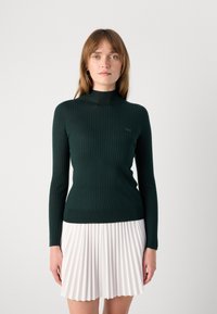 Pull à col roulé côtelé vert foncé avec des manches longues et une silhouette ajustée, associé à une jupe plissée blanche. Détail du logo Lacoste sur la poitrine.