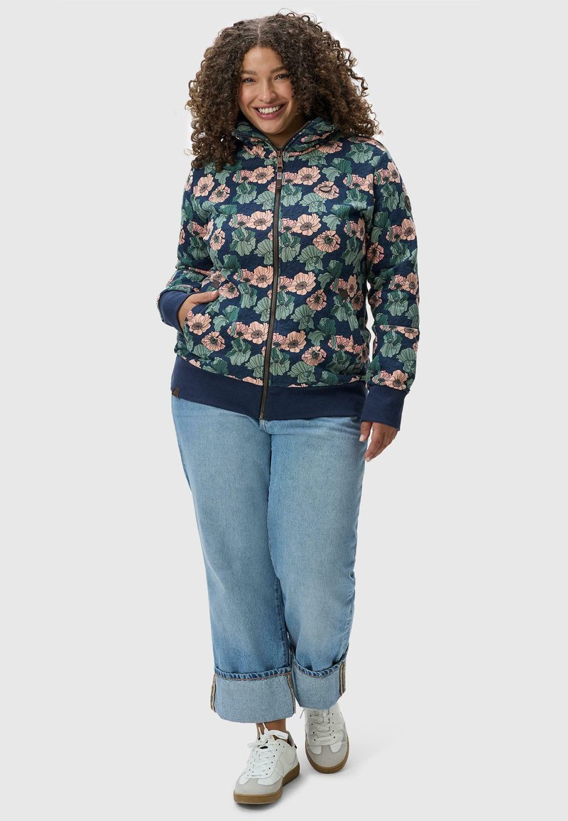 Giacca a zip floreale con design blu scuro e rosa, dotata di cappuccio e polsini a coste, abbinata a jeans a gamba larga azzurri e sneakers bianche.