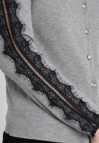Grå cardigan med svart spetsdetalj längs ärmarna, prydd med silverknappar och en ribbad fåll. Spetsen har blommönster med fransdetaljer.
