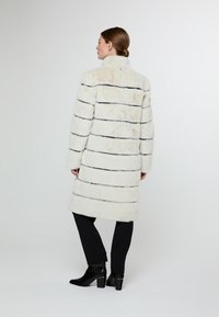 Bílý kabát z umělé kožešiny s černými vodorovnými pruhy, vysokým límcem a uvolněným střihem, doplněný černými zúženými kalhotami a kotníčkovými botami.