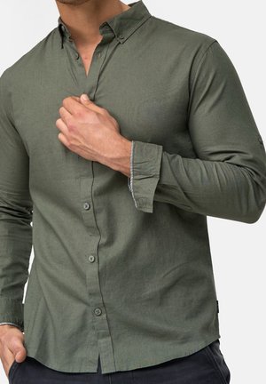 Mężczyzna poprawiający kołnierz oliwkowo-zielonej koszuli na długi rękaw z subtelnym czarno-białym detalem na wewnętrznych mankietach.