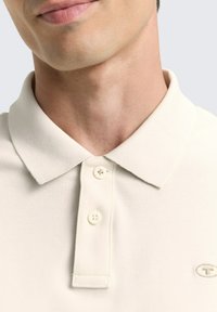 Beige Poloshirt aus strukturiertem Stoff, mit klassischem Kragen, drei Knöpfen und kleinem Logo auf der Brust. Nahaufnahme des Ausschnitts.
