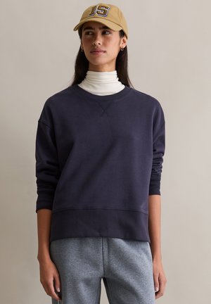 Marineblauwe sweatshirt met geribbelde manchetten en zoom, voorzien van een ronde hals. Gedragen over een witte coltrui, gecombineerd met grijze broek.